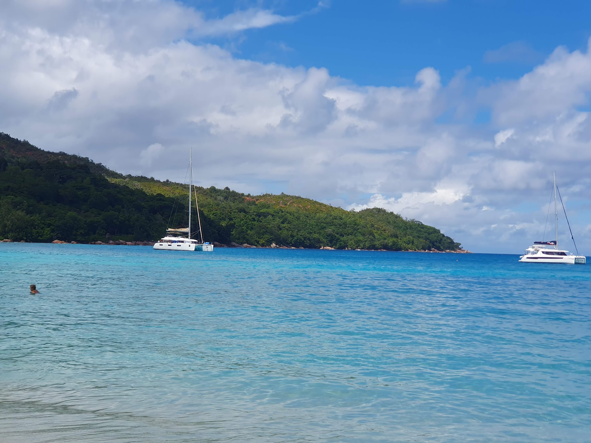 Türkisfarbene Bucht mit zwei vor Anker liegenden weißen Katamaranen, bewaldete Insel im Hintergrund, Schwimmer im Vordergrund.