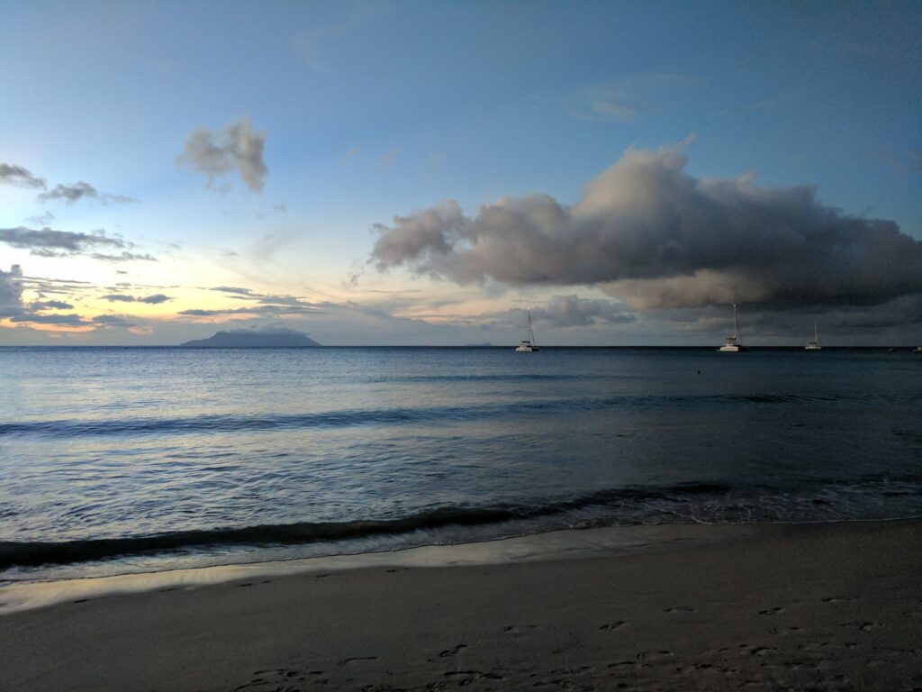 Ruhiger Strand bei Dämmerung; sanfte Wellen, drei Segelboote vor ferne Insel unter wolkigem Himmel.