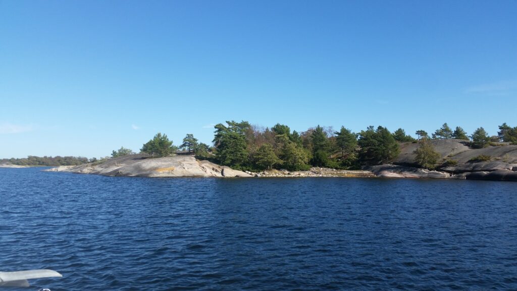 Kleine felsige Insel mit Kiefern, umgeben von tiefblauem Wasser unter wolkenlosem, hellblauem Himmel.