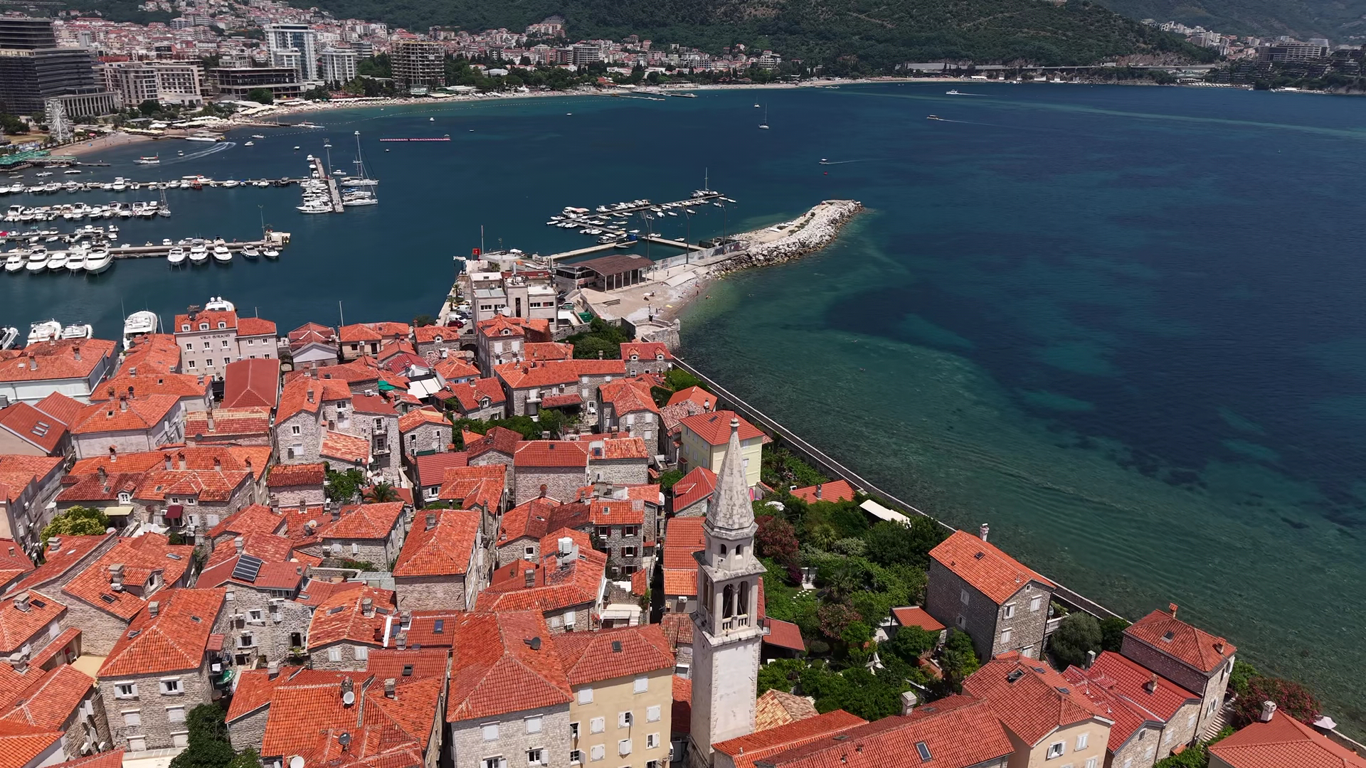 Luftaufnahme einer Altstadt mit roten Ziegeldächern auf einer Landzunge, angrenzender Yachthafen im türkisblauen Meer.