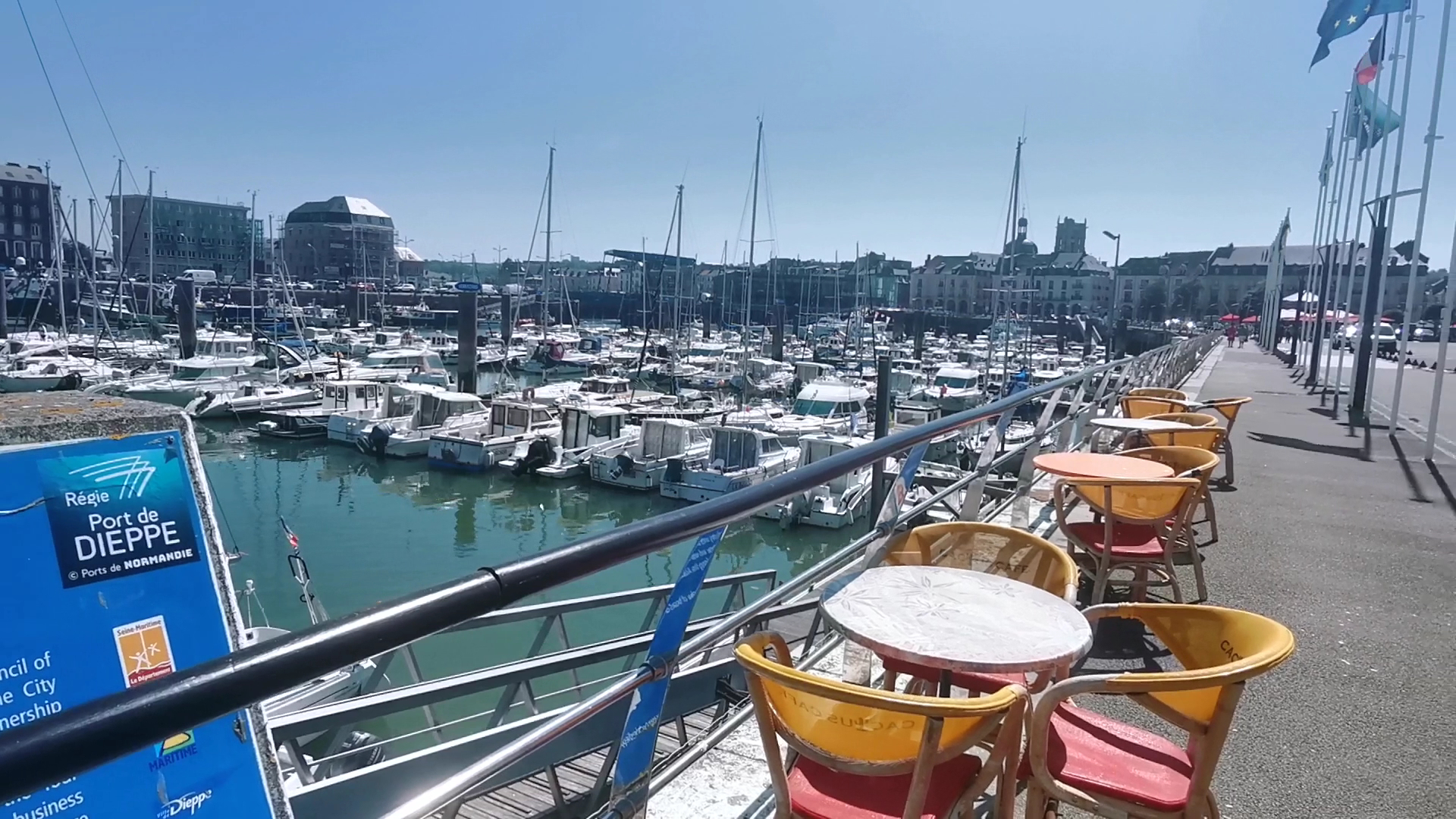 Sonniges Hafenpanorama in Dieppe; Caféterrasse mit roten und gelben Stühlen blickt auf dicht ankernde Boote.