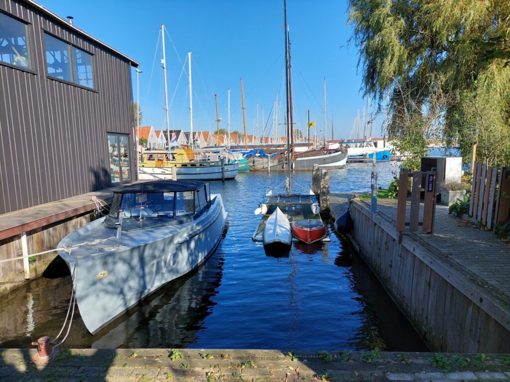 Kleiner Hafen: mehrere Segel- und Motorboote vertäut zwischen Holzstegen, rote Dachhäuser im Hintergrund, blauer Himmel