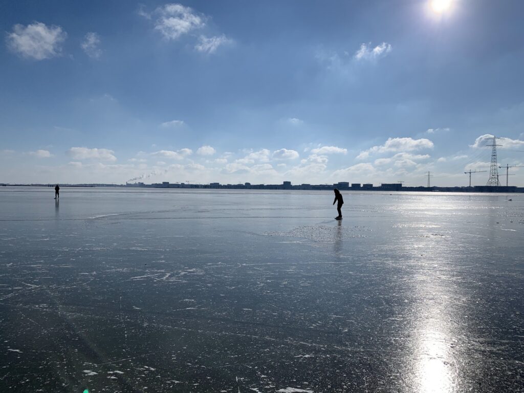 Weitläufiger zugefrorener See mit zwei Eisläufern; helle Wintersonne spiegelt sich, ferne Stadtsilhouette am Horizont.