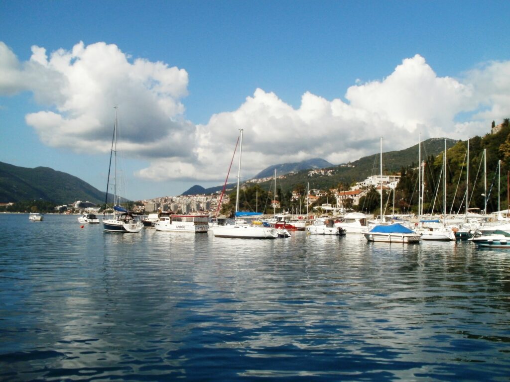 Ruhiger Hafen mit zahlreichen Segel- und Motorbooten; dahinter Küstenstadt, grüne Berge und blauer Himmel mit Wolken.