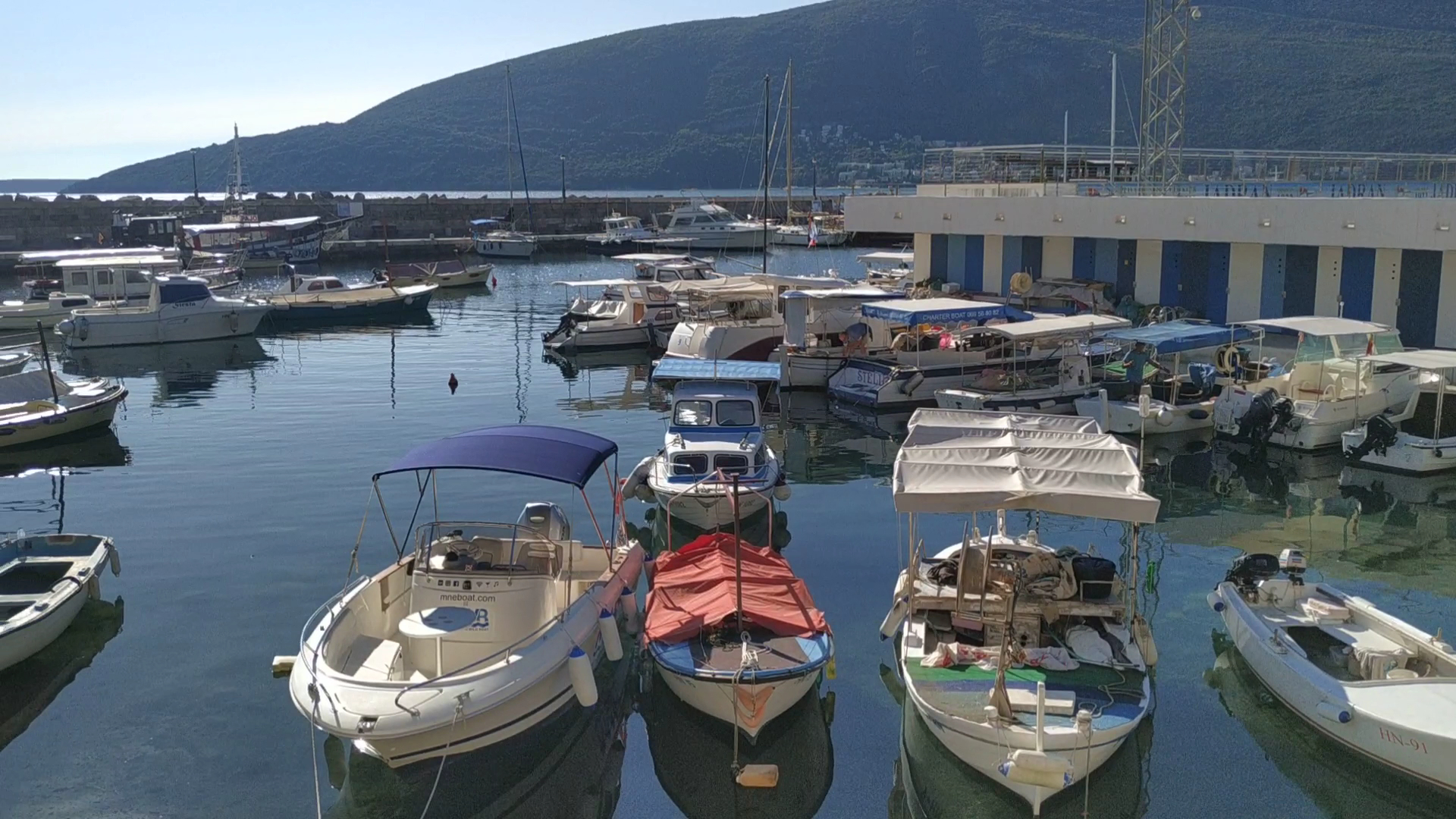Ruhiger Yachthafen: kleine Motor- und Segelboote liegen dicht aneinander, dahinter Meer und grüner Berg.