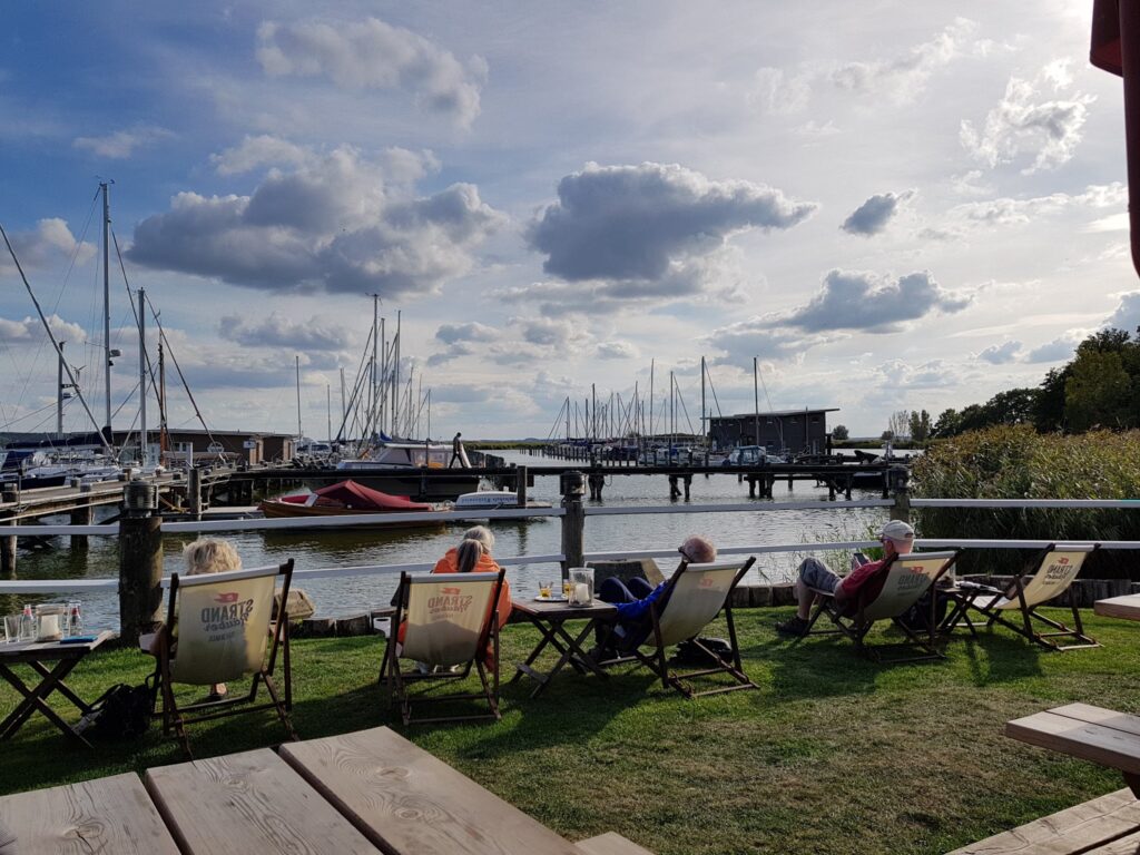 Mehrere Personen entspannen in Liegestühlen am Hafen; im Hintergrund Segelboote, Stege und leichter Wolkenhimmel.