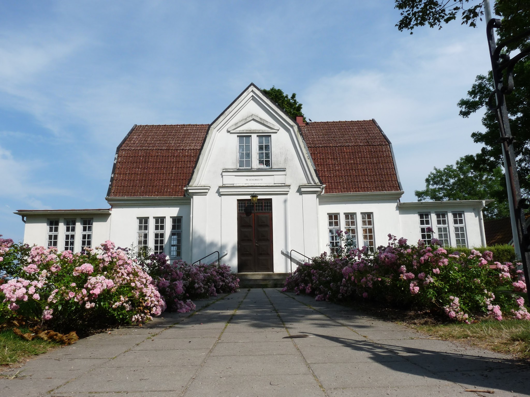Weißes Haus mit rotem Giebeldach; zentraler Holzeingang, Weg beidseitig von üppigen rosa Rosen gesäumt.