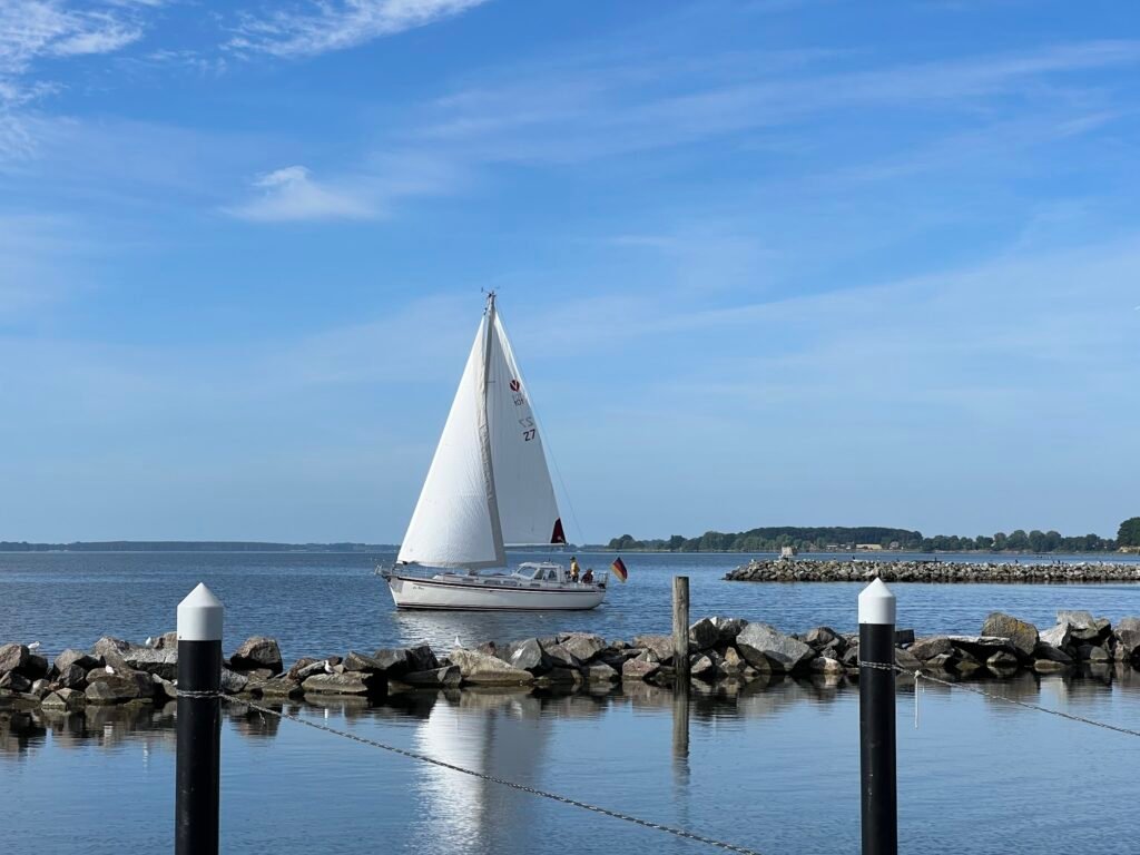 Segelboot mit deutscher Flagge gleitet vor Steinmole auf ruhiger See; blauer Himmel, zwei schwarze Dalben im Vordergrund.