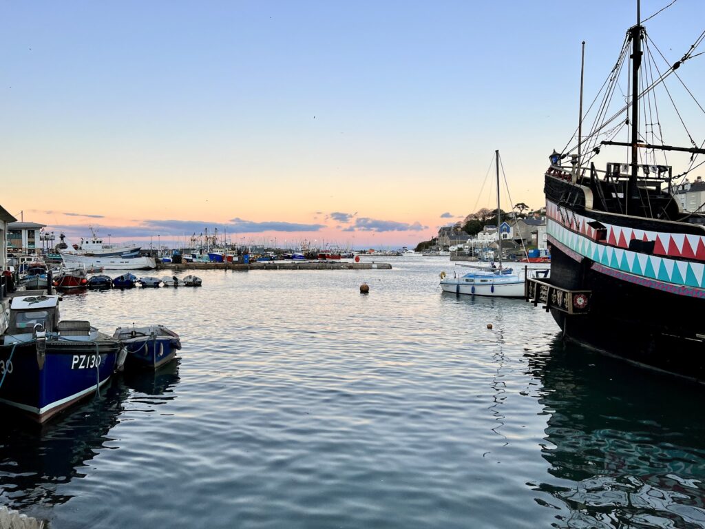 Hafen im Abendlicht, kleine Fischerboote und großes schwarz-buntes Segelschiff vor rosa-blauem Himmel.