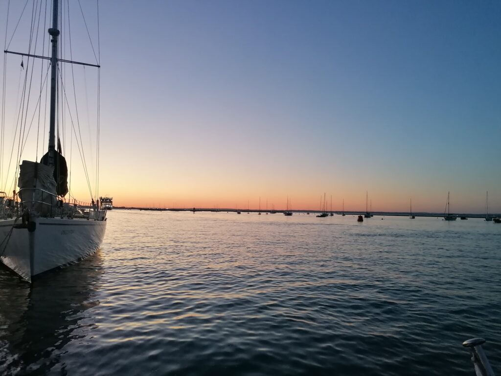 Segelboot im Vordergrund vor ruhigem Wasser; am Horizont liegen weitere Boote unter farbigem Sonnenuntergangshimmel.