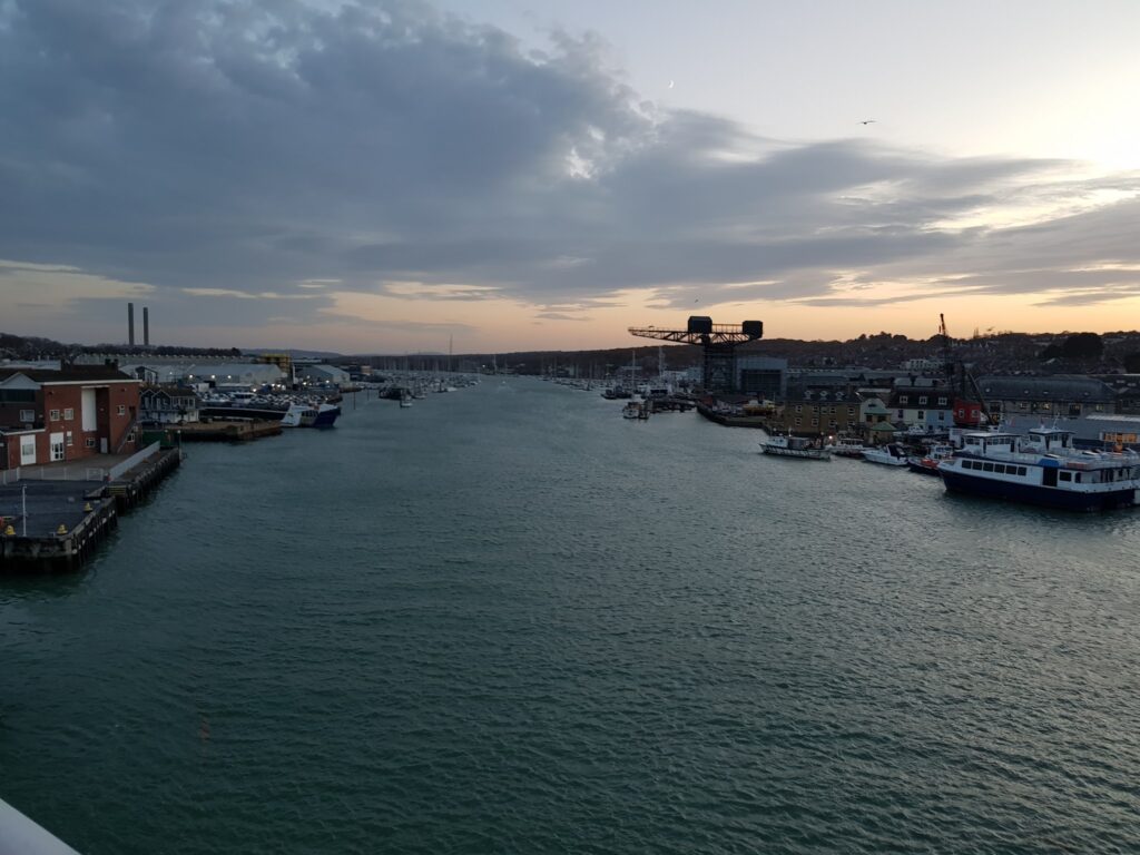 Hafenkanal bei Dämmerung, Boote an beiden Ufern vertäut, Lagerhallen, Kran, ferne Schornsteine, ruhiges Wasser, Sichelmond.