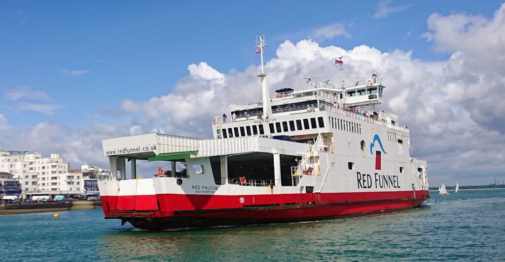 Weiße Autofähre „Red Funnel“ mit rotem Rumpf fährt im Hafen, blauer Himmel und Küstenhäuser im Hintergrund.