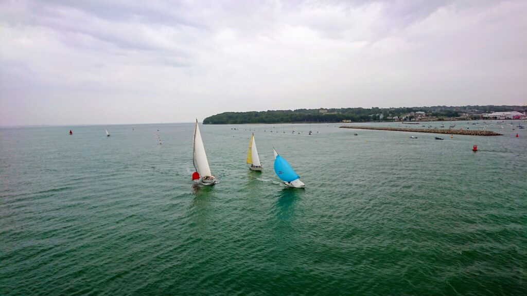 Drei Segelboote mit weißen, gelben und blauen Segeln kreuzen auf ruhiger See vor einer bewaldeten Küste bei Wolkenhimmel.