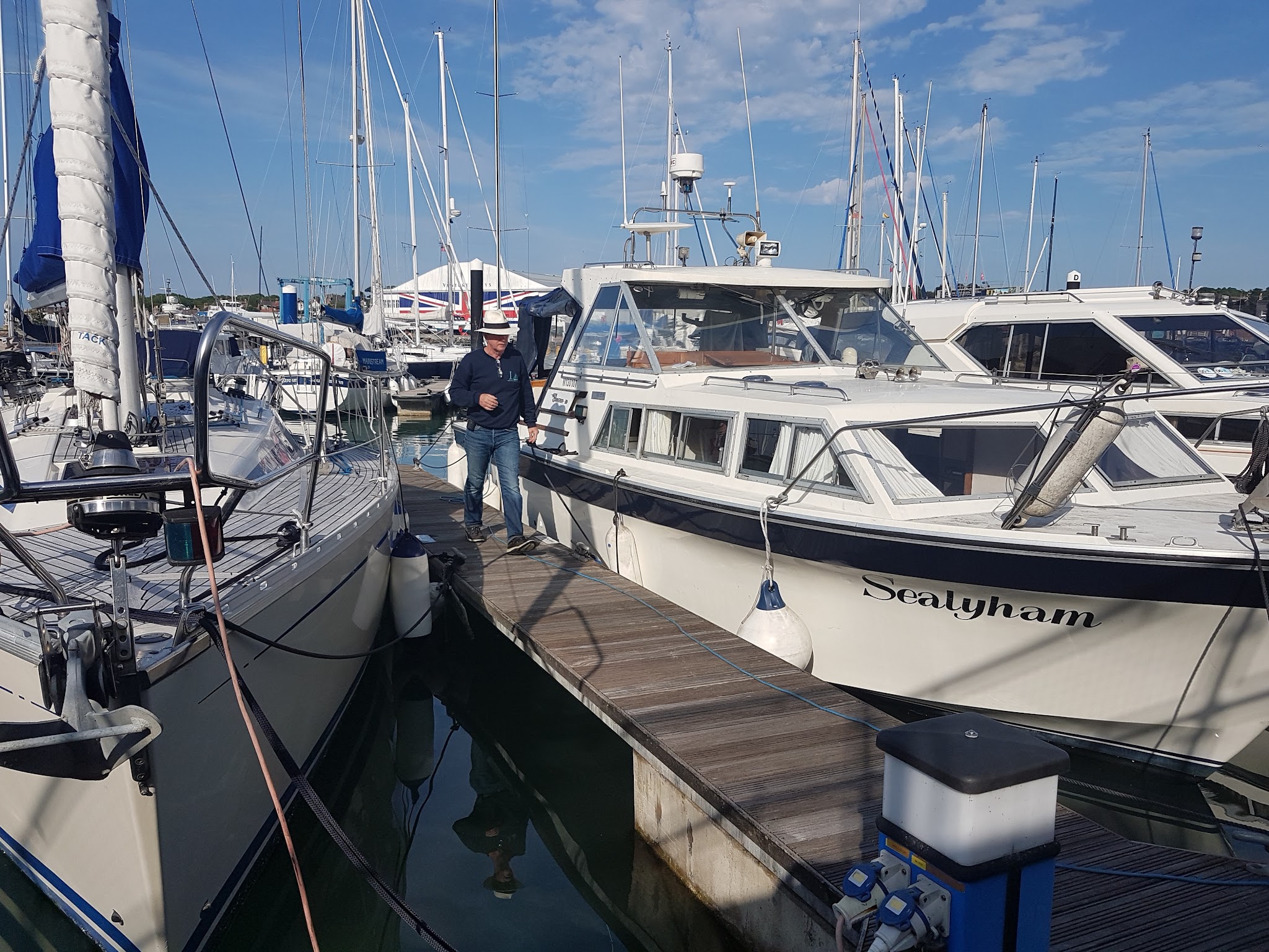Mann mit Hut geht auf Steg zwischen Segelyacht und Motorboot „Sealyham“ im Yachthafen unter blauem Himmel.