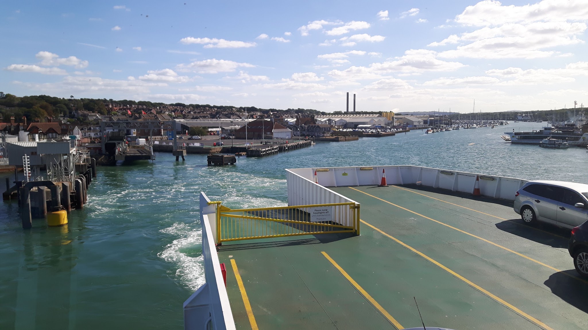Fähre verlässt Hafen; leeres Autodeck vorn, Gischt im Wasser, Stadthafen mit Booten und Häusern im Hintergrund