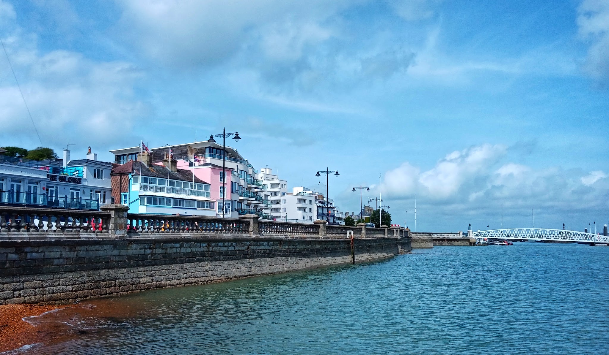 Pastellfarbene Küstenhäuser entlang einer Steinpromenade, ruhiges Meer und weiße Brücke unter blauem Himmel.
