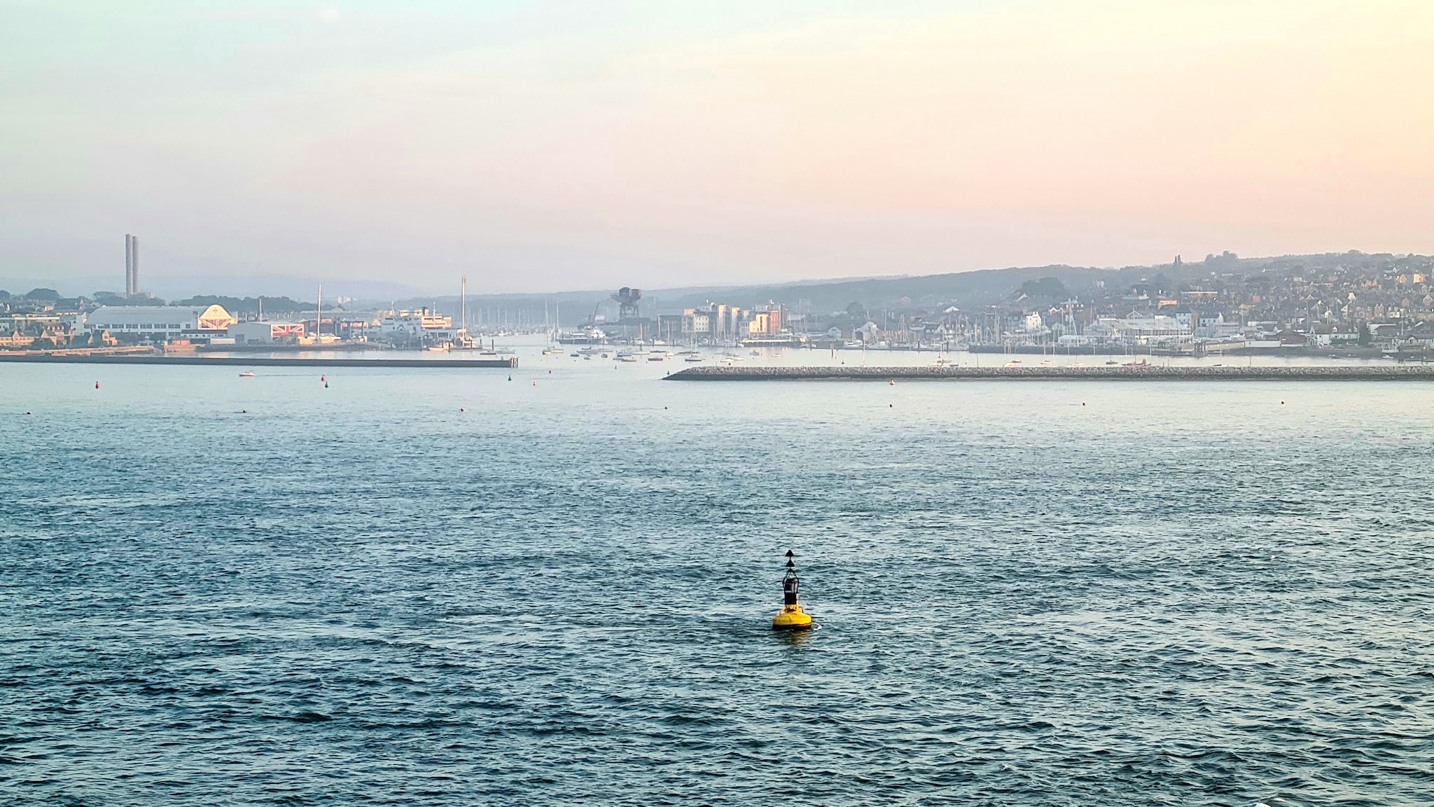 Gelbe Boje treibt im ruhigen Wasser vor einem morgendlichen Hafen mit Stadt und Booten unter pastellfarbenem Himmel