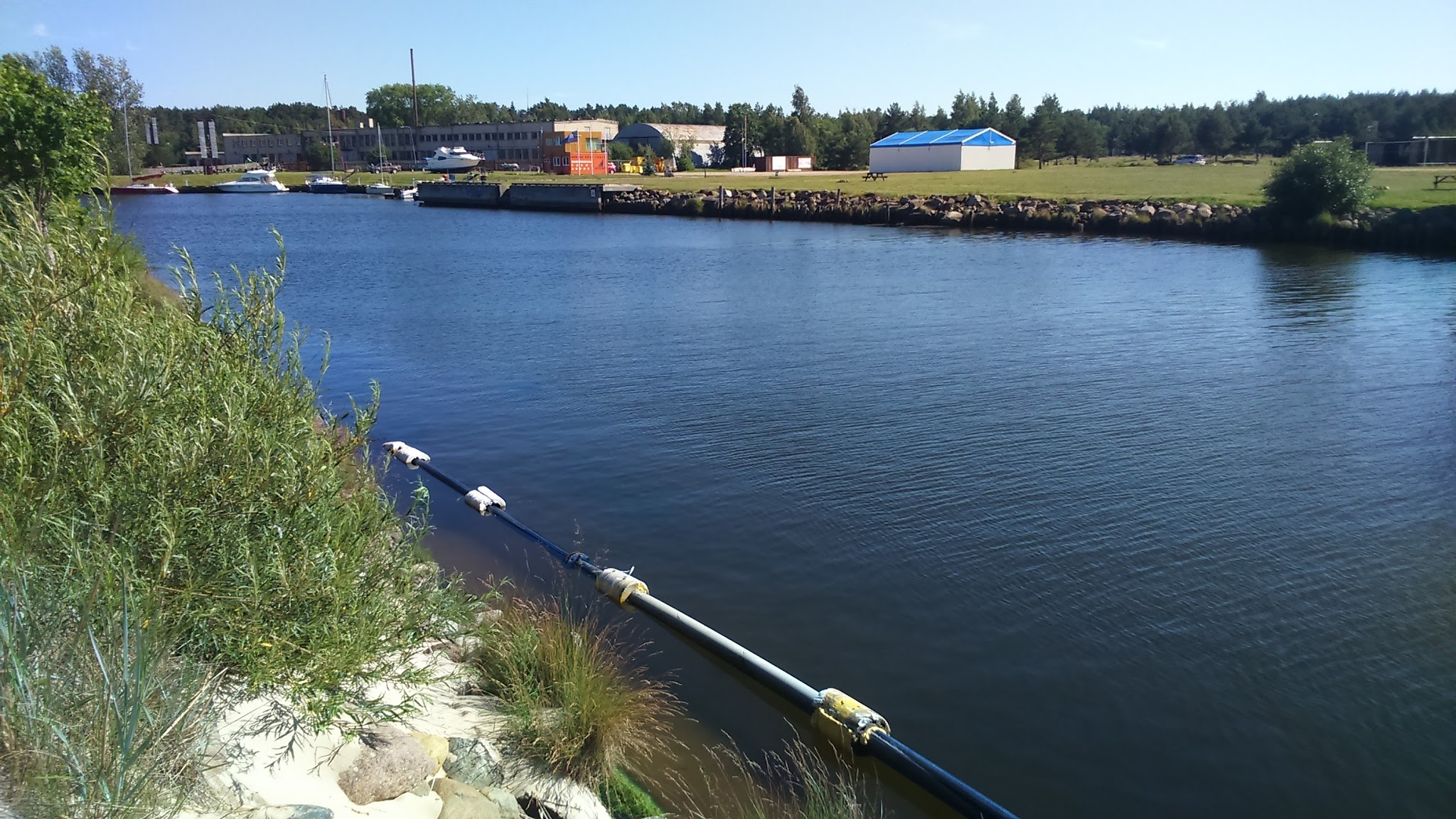 Ruhiger Kanal mit schwimmender Rohrleitung; gegenüber Anlegestelle mit Booten, Wiese und niedrigen Gebäuden.
