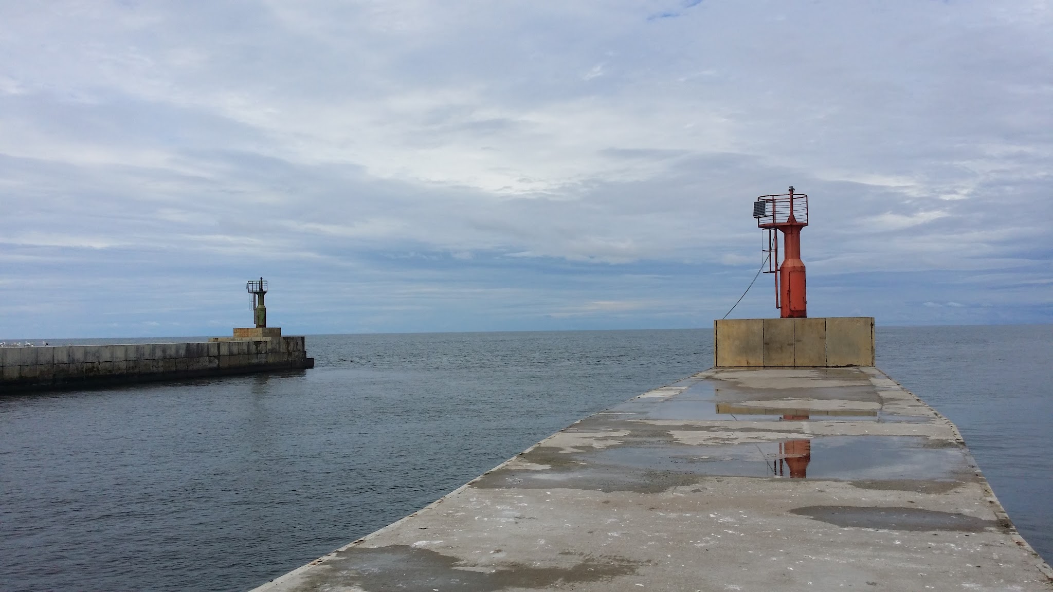 Betonpier mit rotem Leuchtfeuer im Vordergrund, gegenüber grüne Bake; ruhiges Meer unter bewölktem Himmel
