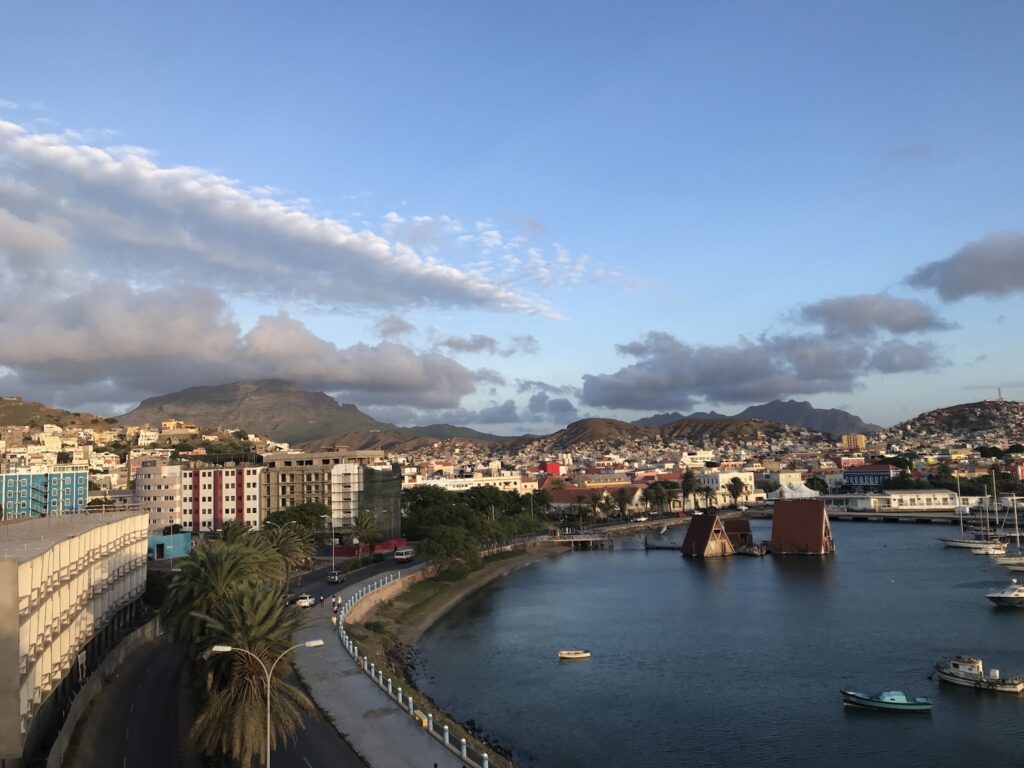 Bunte Küstenstadt vor grünen Bergen; Palmallee am Wasser, Boote und zwei schwimmende Häuser mit Spitzdach im Hafen.