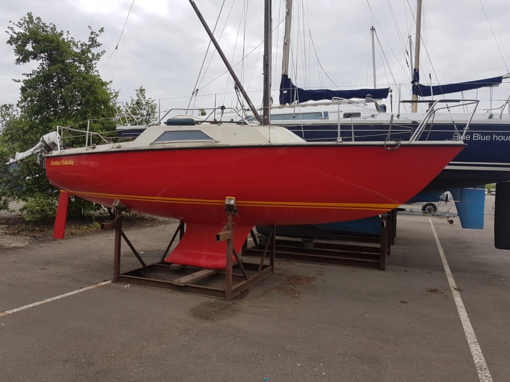 Rotes Segelboot steht auf Böcken im Hafenlager, Mast hoch, blaues Boot im Hintergrund.
