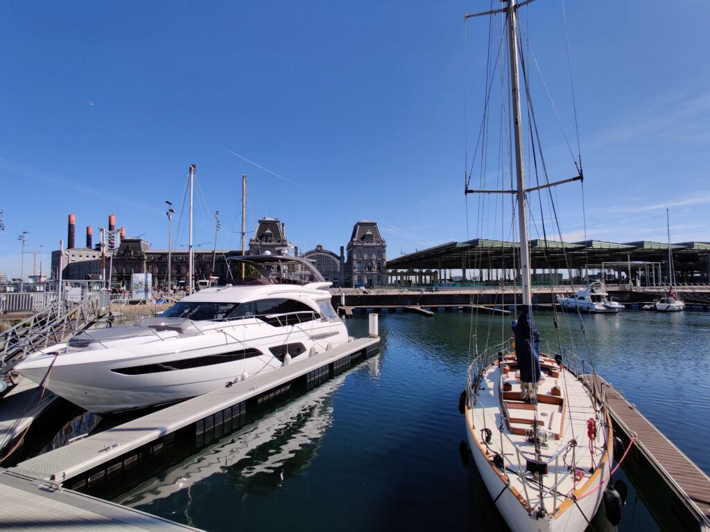 Sonniger Hafen: moderne Motoryacht und Segelboot liegen am Steg vor historischen Backsteinbauten und blauem Himmel.