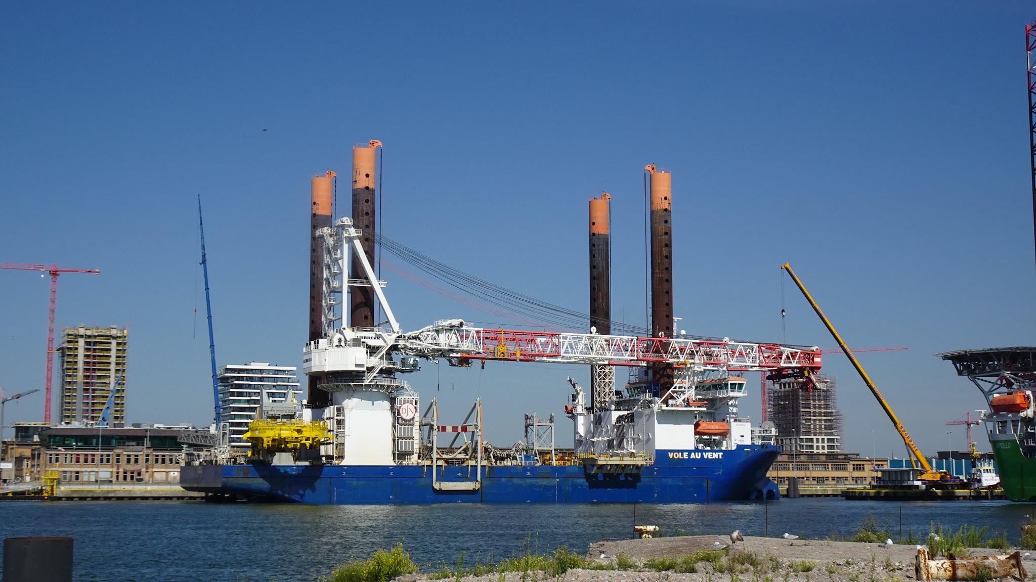 Großes Offshore-Installationsschiff mit vier hohen Stelzen und langem Kranarm liegt im Hafen vor Baustellenkulisse.