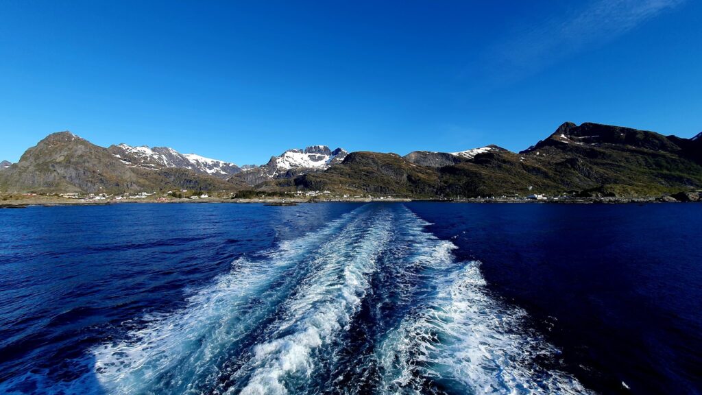 Fährspur im tiefblauen Meer, führt zu schneebedeckten norwegischen Fjordbergen unter wolkenlosem Himmel.