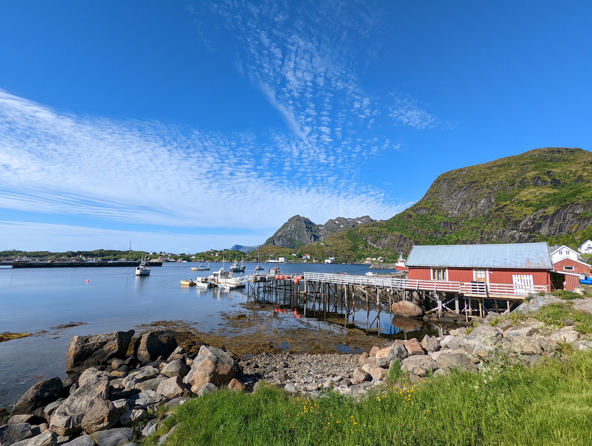 Ruhiger Fjord mit kleinen Fischerbooten, rotem Pfahlboathaus, grünen Bergen und blauem Himmel mit Schleierwolken.