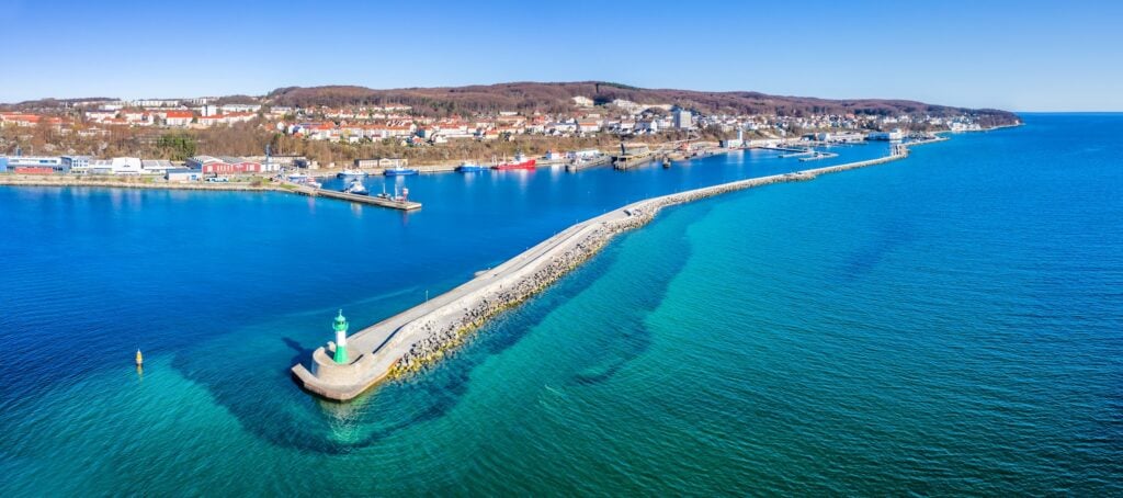 Langer Wellenbrecher mit grün-weißem Leuchtturm ragt ins türkisblaue Meer vor Küstenstadt unter klarem Himmel.
