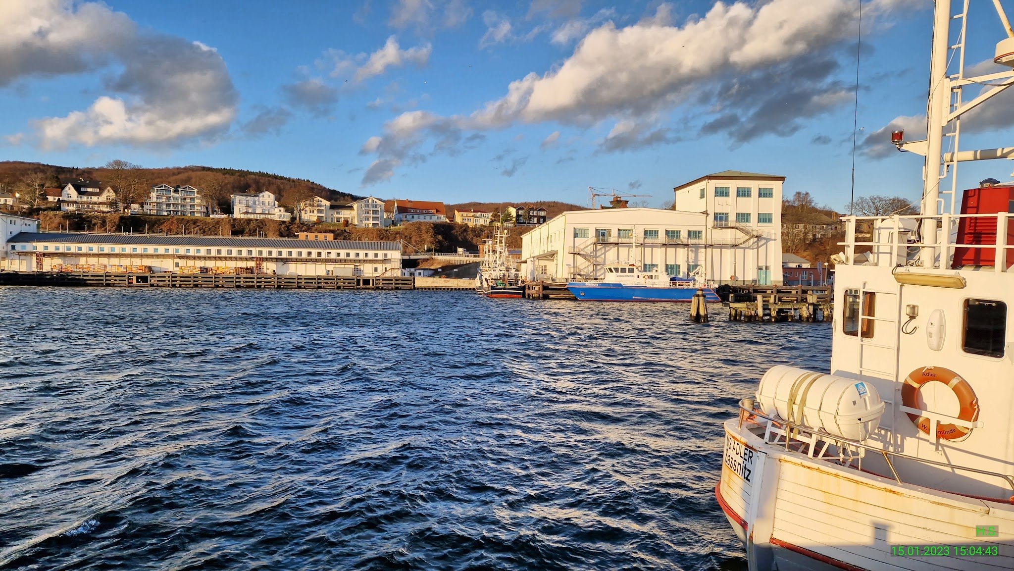 Sonniger Hafen: Vorn weißes Ausflugsschiff, dahinter welliges Wasser, Lagerhallen und Hügel mit Häusern.