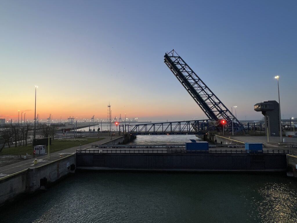 Geöffnete Klappbrücke über Schleuse, dahinter Hafenkräne im Morgendämmerlicht, rote Ampel signalisiert Stopp.