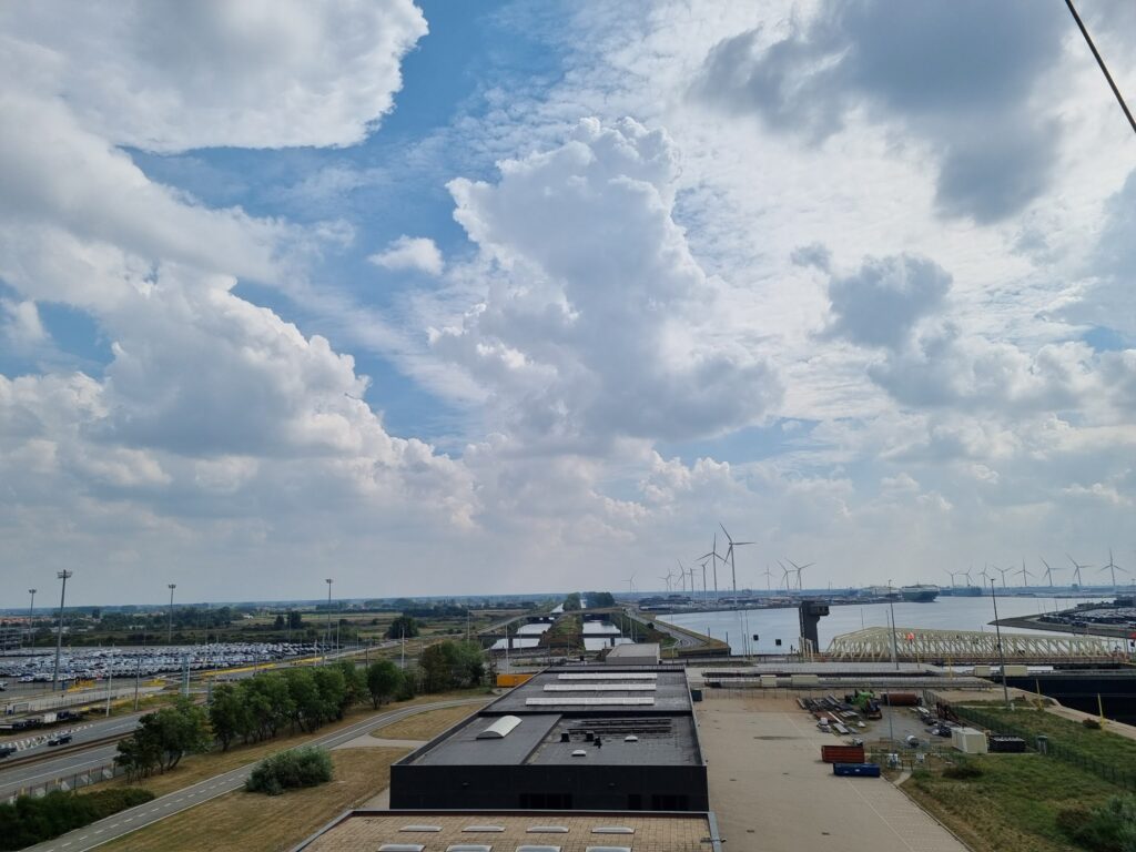 Weitläufiger Hafen mit Hallendächern, Wasserkanal und vielen Windrädern unter dramatischem Wolkenhimmel.