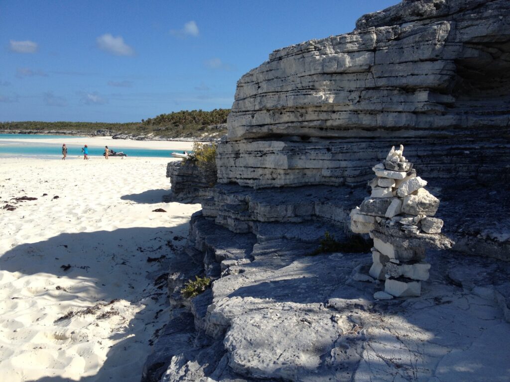 Kalksteinfelsen mit Steinmännchen am weißen Strand; im Hintergrund Spaziergänger und türkisblaues Meer.