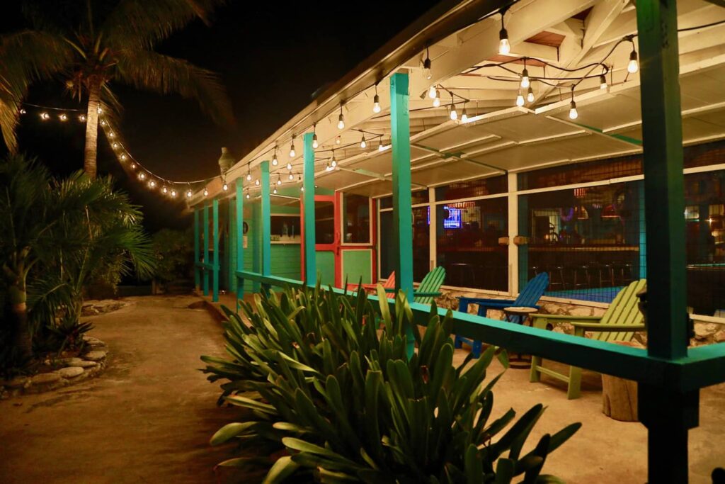 Strandbar bei Nacht, Lichterkette über türkisgrüner Veranda, bunte Liegestühle, umgeben von Palmen und Pflanzen.