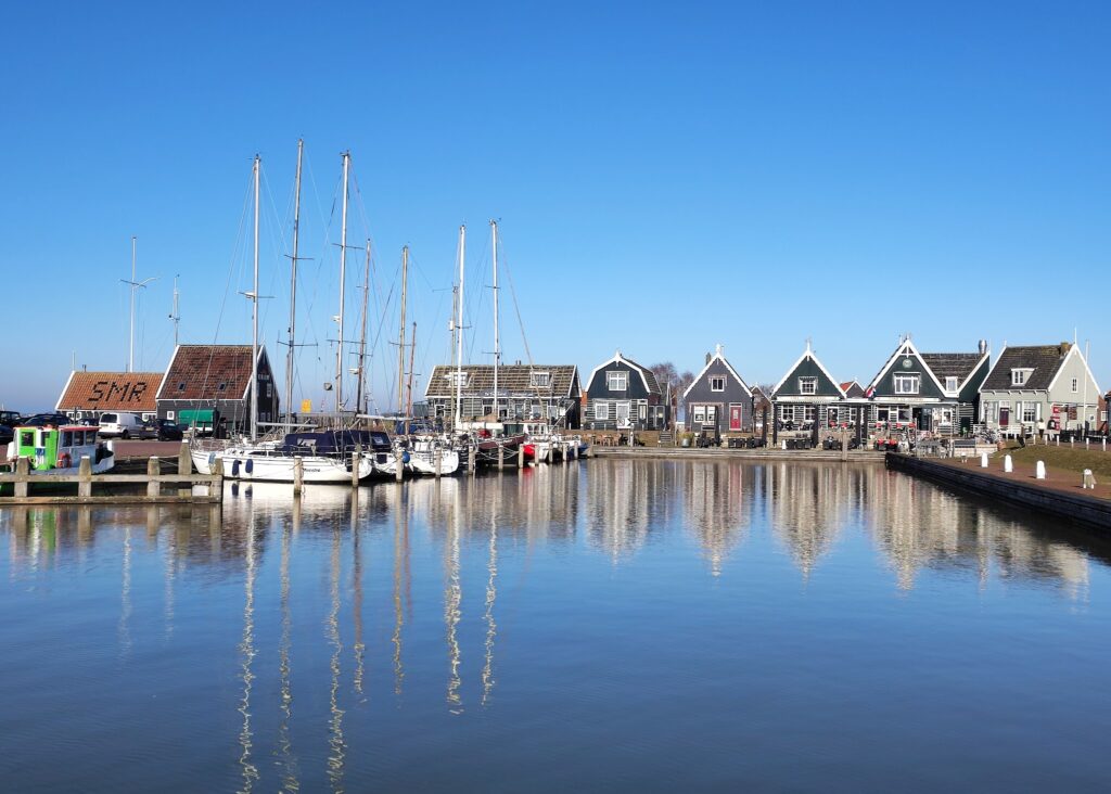 Ruhiger Hafen mit vertäuten Segelbooten, davor spiegeln sich ihre Masten und bunte Holzhäuser im klaren Wasser.