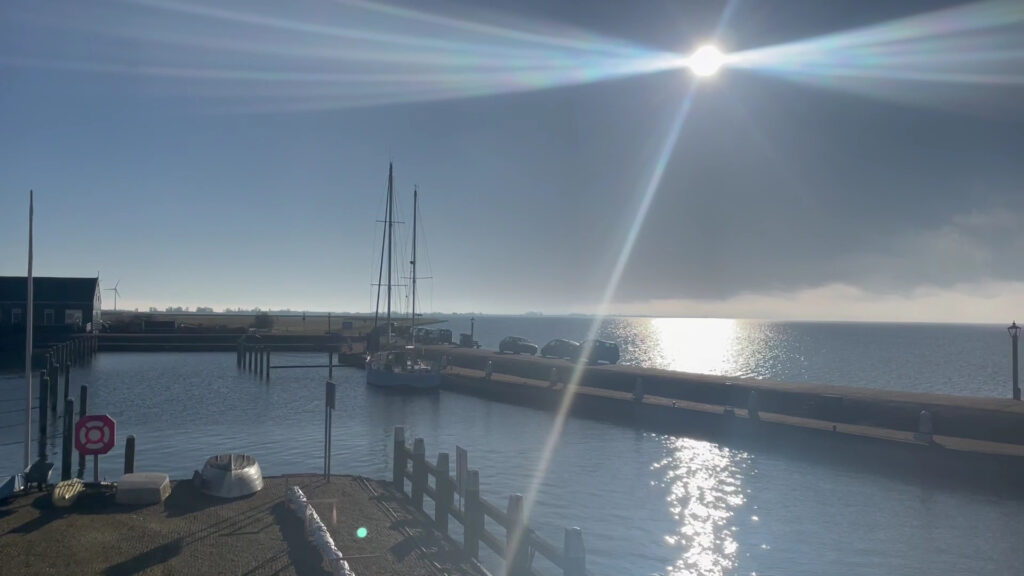Heller Morgen im kleinen Hafen: Segelboot am Steg, Sonne glitzert im ruhigen Wasser, wenige Autos auf der Mole.
