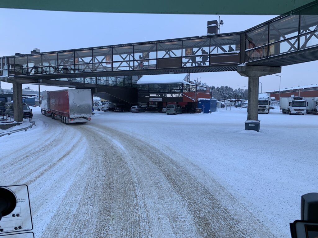 Verschneiter Speditionshof; Lkw fährt unter gläsernem Fußgängersteg, weitere Lkw und Hallen im Hintergrund.