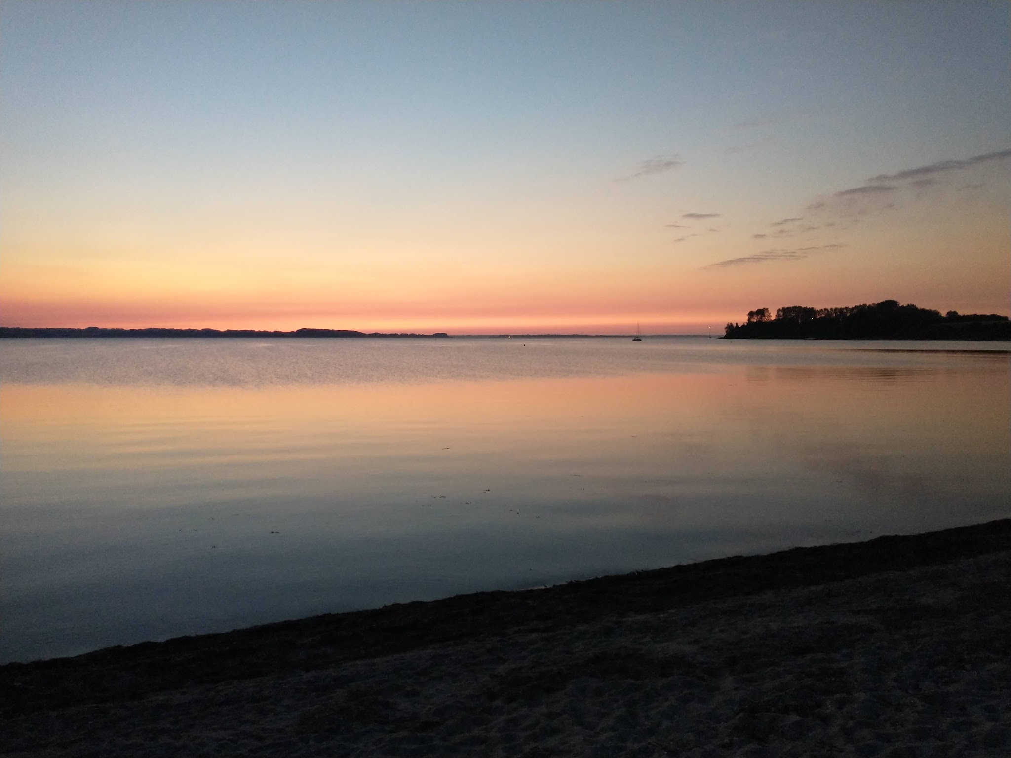 Ruhiger Strand bei Sonnenuntergang, glattes Wasser spiegelt orange-blauen Himmel; ferne Landzunge, kleines Segelboot