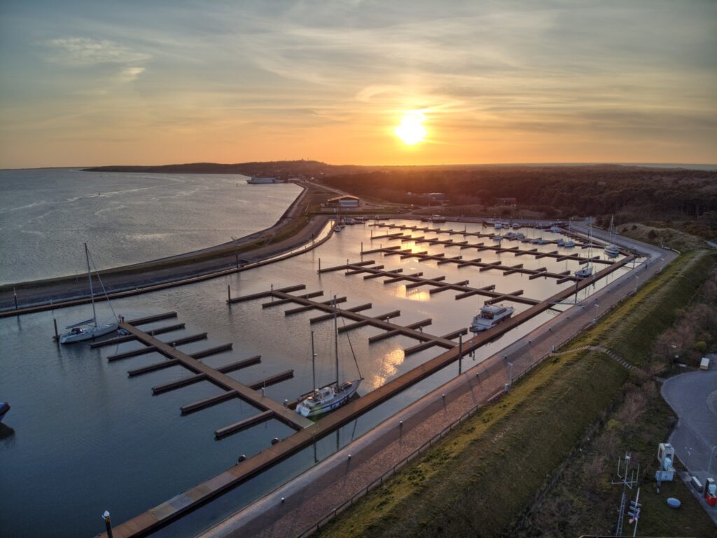 Luftaufnahme eines Hafens bei Sonnenuntergang, leere Stege spiegeln sich im Wasser, wenige Segelboote festgemacht.