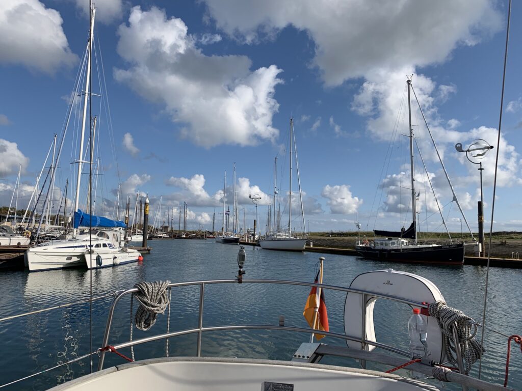 Ruhiger Yachthafen mit vielen Segelbooten, Blick vom Heck eines Boots auf blaues Wasser unter lockeren Wolken.