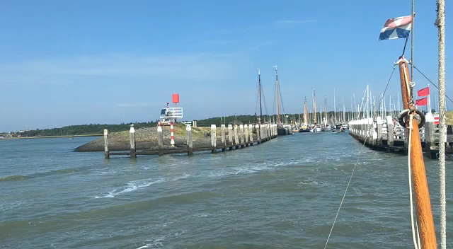 Schmale Hafeneinfahrt zwischen Pfählen führt zu zahlreichen Segelbooten; ruhiges Wasser, klarer Himmel.