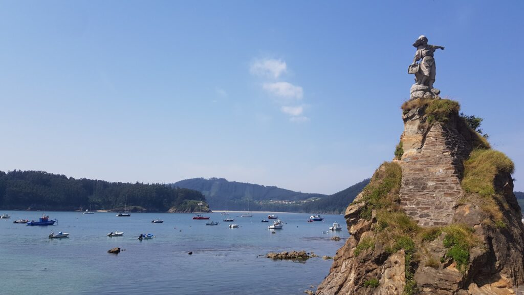 Statue auf felsiger Klippe zeigt aufs Meer; unten ruhen zahlreiche Boote in ruhiger Bucht unter blauem Himmel.