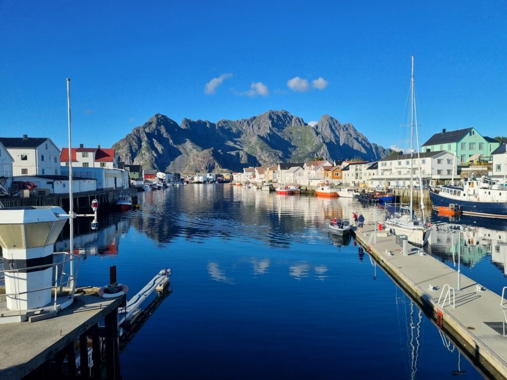 Küstenhafen mit bunten Holzhäusern, Booten und steilen Bergen, die sich im ruhigen, klaren Wasser spiegeln.