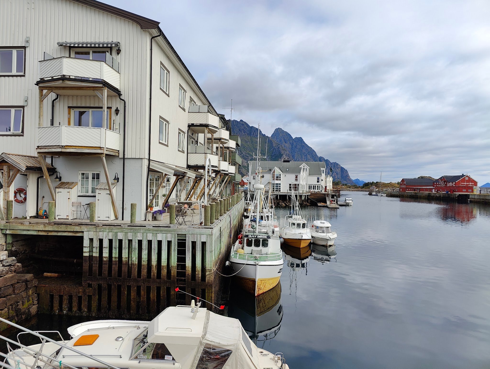 Kleiner Hafen mit weißen Holzhäusern auf Stelzen und mehreren Fischerbooten, dahinter steile Berge unter Wolkenhimmel.