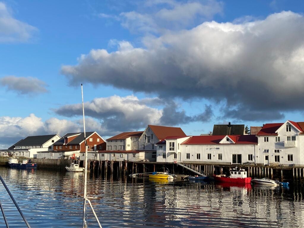 Kleine Hafenhäuser mit roten Dächern spiegeln sich im ruhigen Wasser; bunte Boote liegen am Kai unter Wolkenhimmel.