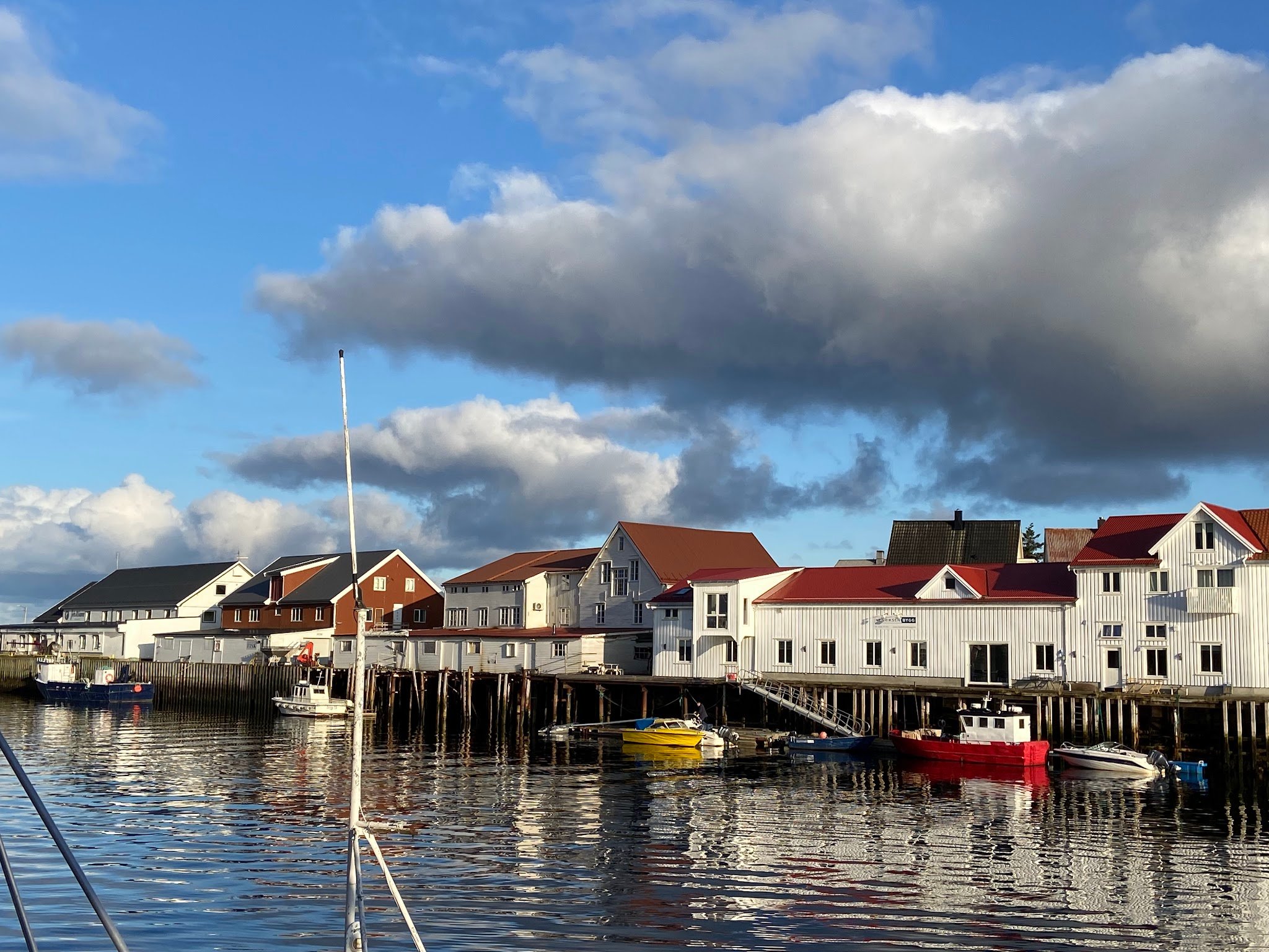 Kleine Hafenhäuser mit roten Dächern spiegeln sich im ruhigen Wasser; bunte Boote liegen am Kai unter Wolkenhimmel.
