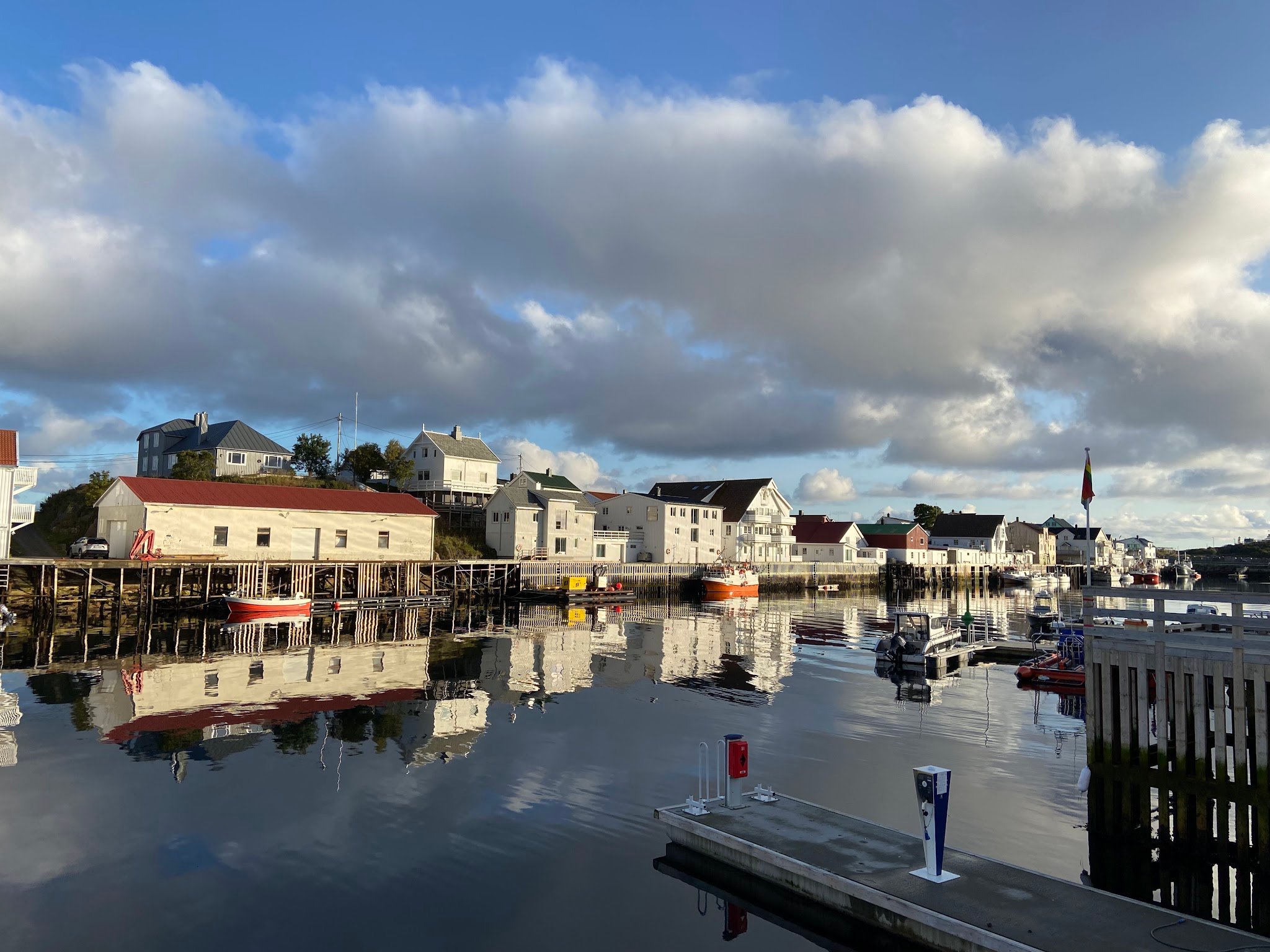Ruhiger Hafen: weiße Holzhäuser auf Stelzen spiegeln sich im Wasser, kleine Boote vertäut unter wolkigem Himmel.