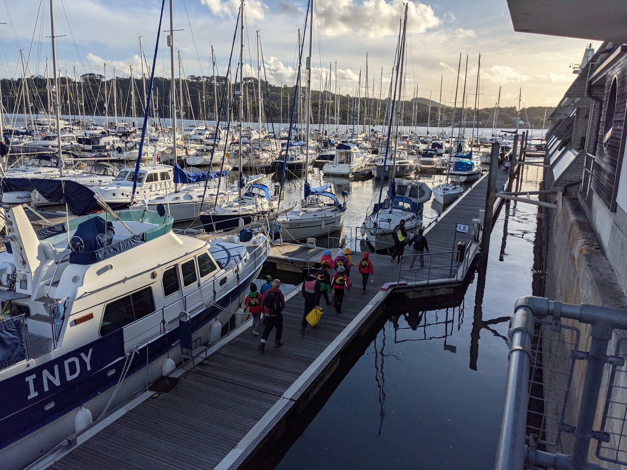 Gruppe in bunten Schwimmwesten geht auf Steg zwischen dicht liegenden Segelbooten im Yachthafen bei tiefem Sonnenlicht.