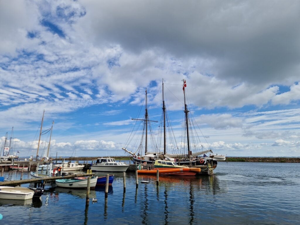 Zweimast-Segelschiff und kleine Boote liegen im sonnigen Hafen; ruhiges Wasser spiegelt Wolken und blauen Himmel